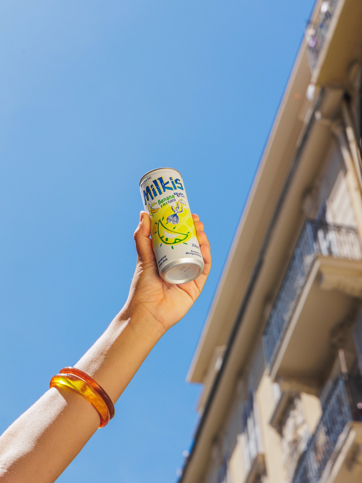 Canette de Milkis levée vers le ciel bleu par une main aux bracelets colorés