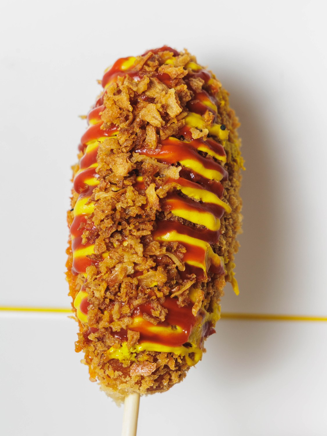 Rice Dog Chungchun croustillant, nappé de ketchup et moutarde, tenu sur son pic