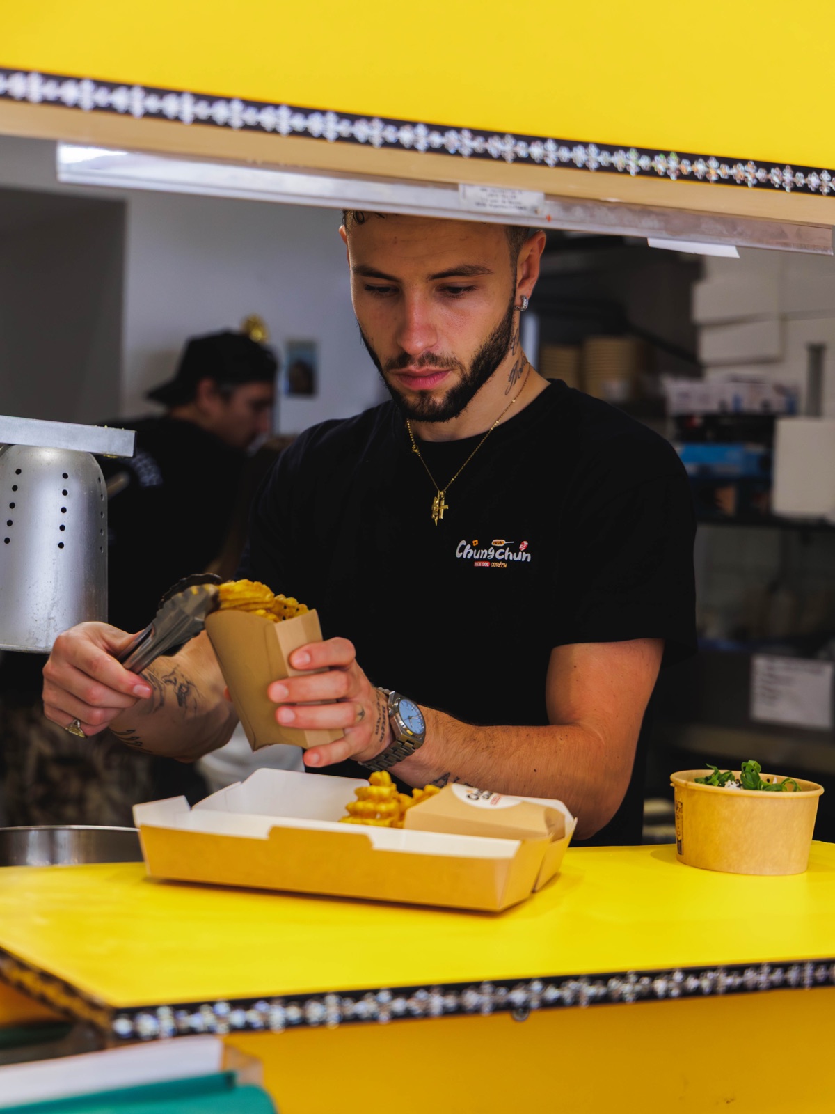 Rice Dog Master Chungchun assemblant un Rice Dog derrière le comptoir jaune