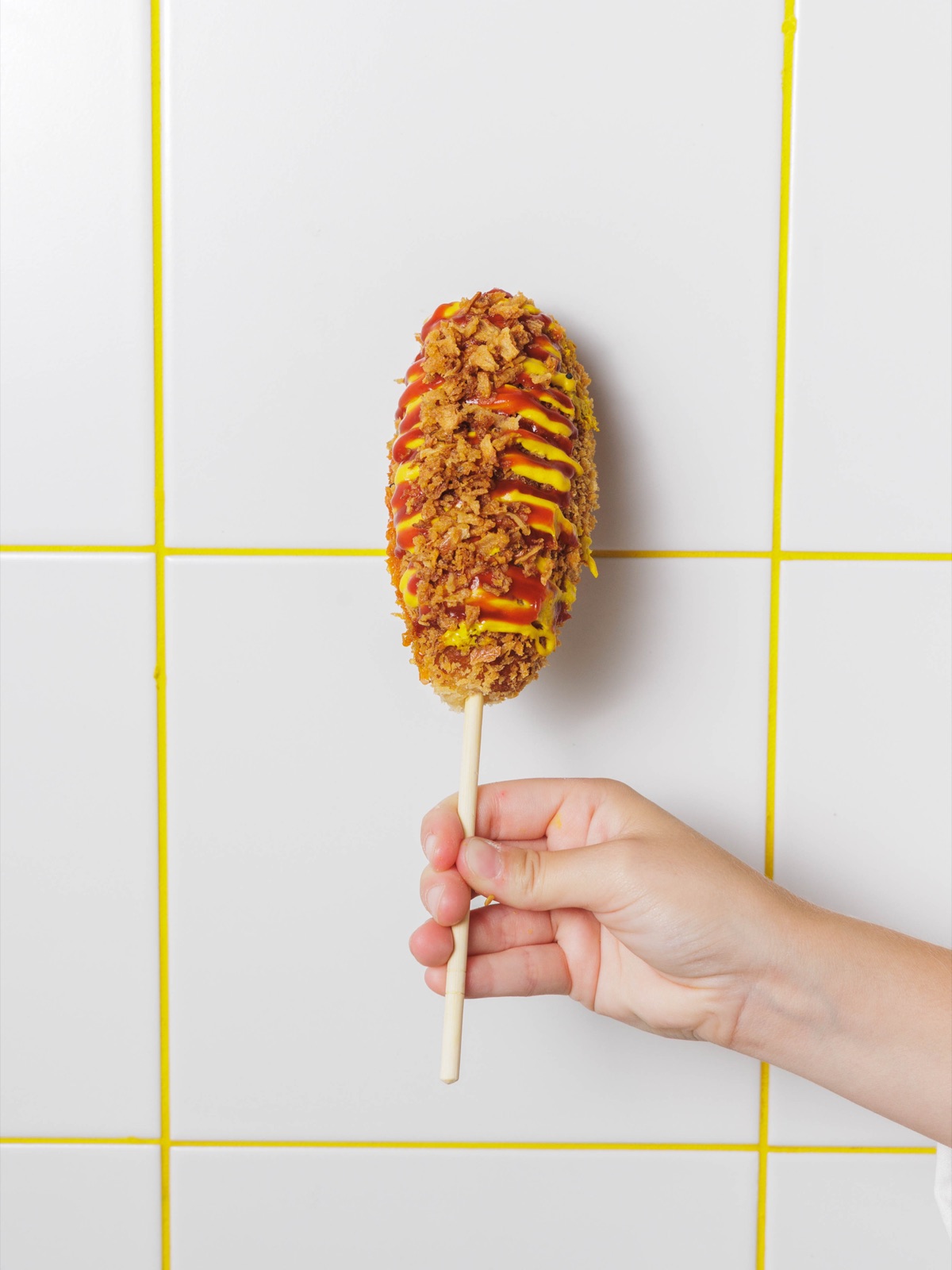 Rice Dog croustillant nappé de ketchup et moutarde, tenu devant le mur carrelé jaune de Chungchun